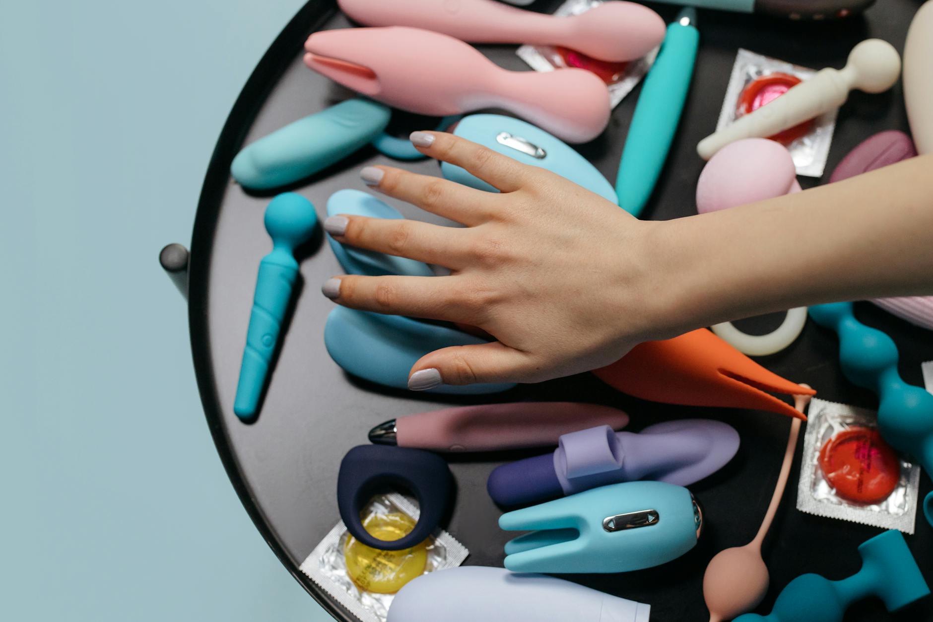 Uma mão alcançando uma variedade de brinquedos sexuais coloridos dispostos em uma mesa