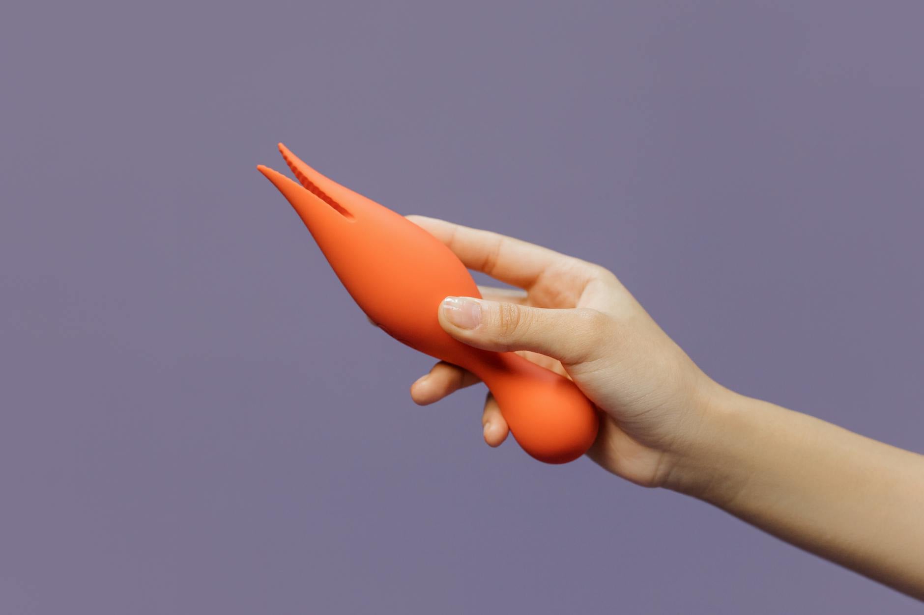 Mão segurando um vibrador de limão laranja contra fundo minimalista roxo, exibindo sensualidade moderna