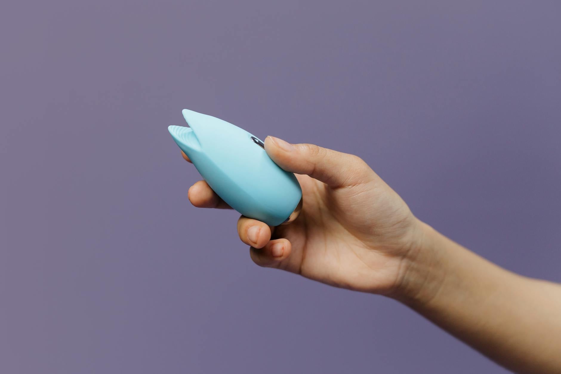 Um vibrador de silicone azul segurado em mão contra um fundo roxo sólido, promovendo amor-próprio e sexualidade